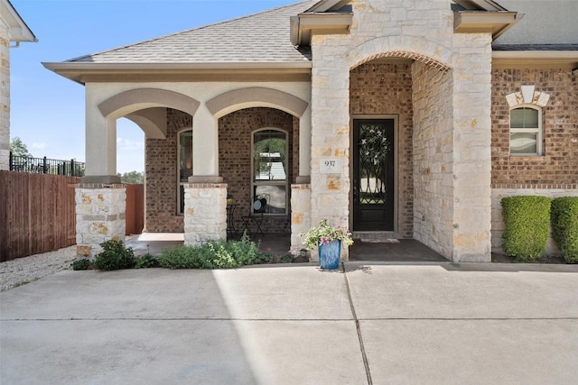 entrance to property featuring brick siding, a shingled roof, fence, and stone siding