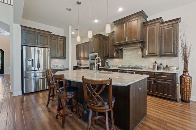 kitchen with appliances with stainless steel finishes, dark wood-style flooring, dark brown cabinetry, tasteful backsplash, and a kitchen island with sink