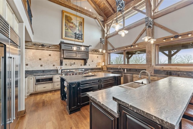 kitchen featuring a center island with sink, built in appliances, dark countertops, a sink, and beamed ceiling