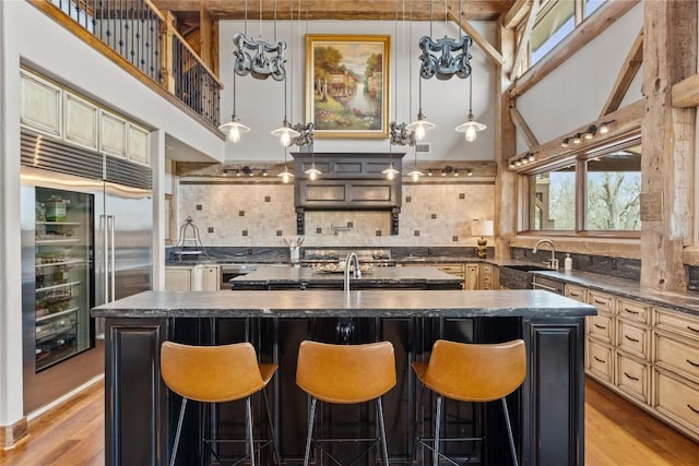 kitchen featuring dark countertops, a sink, a kitchen island with sink, backsplash, and light wood-type flooring