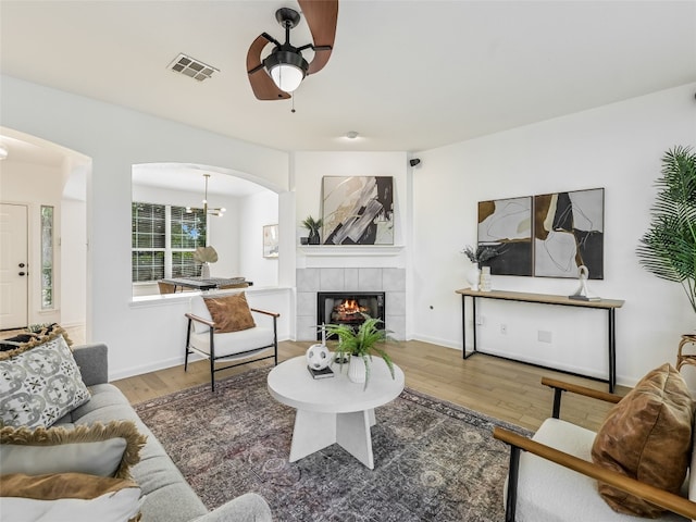 living room featuring arched walkways, ceiling fan, wood finished floors, a tiled fireplace, and a chandelier
