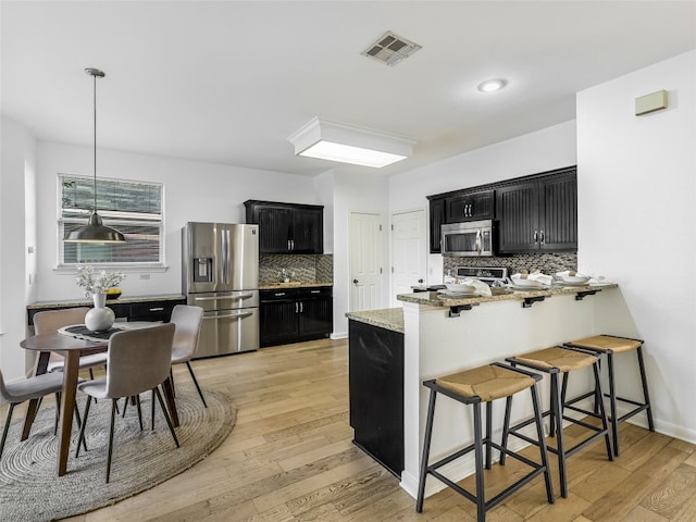kitchen with appliances with stainless steel finishes, dark cabinetry, a kitchen bar, light wood finished floors, and tasteful backsplash