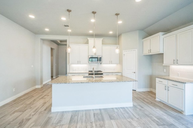 kitchen featuring stainless steel microwave, a kitchen island with sink, tasteful backsplash, light wood finished floors, and light stone countertops