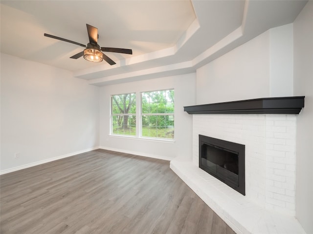 unfurnished living room featuring baseboards, wood finished floors, a ceiling fan, a brick fireplace, and a tray ceiling