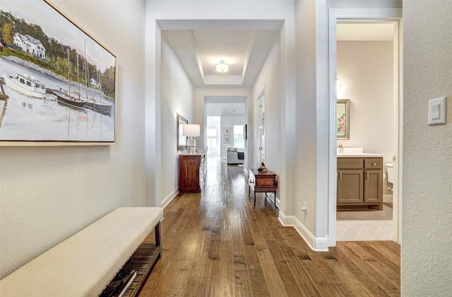corridor featuring wood-type flooring, a raised ceiling, a textured wall, and baseboards