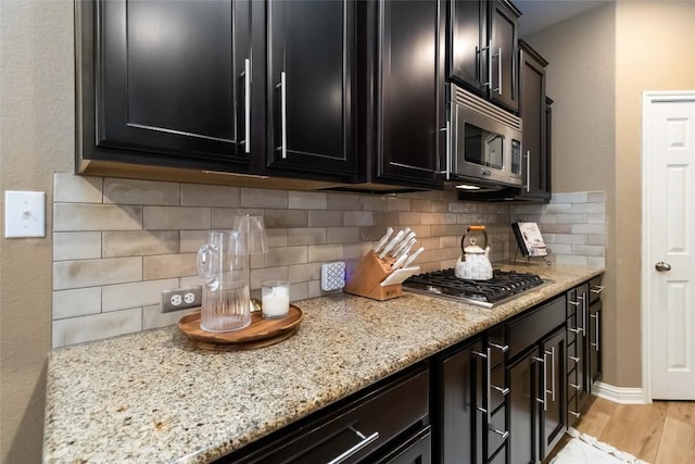 kitchen featuring appliances with stainless steel finishes, dark cabinetry, light wood-type flooring, light stone counters, and decorative backsplash