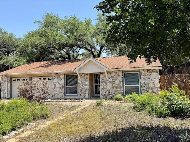 ranch-style home with stone siding, a garage, fence, and roof with shingles