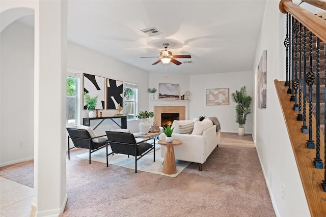 living area featuring ceiling fan, light colored carpet, a warm lit fireplace, and stairs