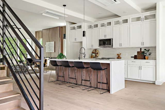 kitchen with stainless steel microwave, backsplash, a sink, a center island with sink, and light wood finished floors