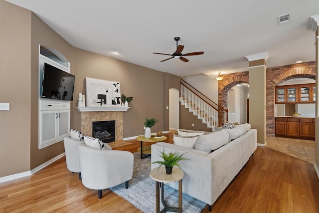 living area with arched walkways, a ceiling fan, a tiled fireplace, and light wood-type flooring