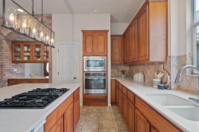kitchen featuring stainless steel appliances, a sink, brown cabinets, light countertops, and glass insert cabinets