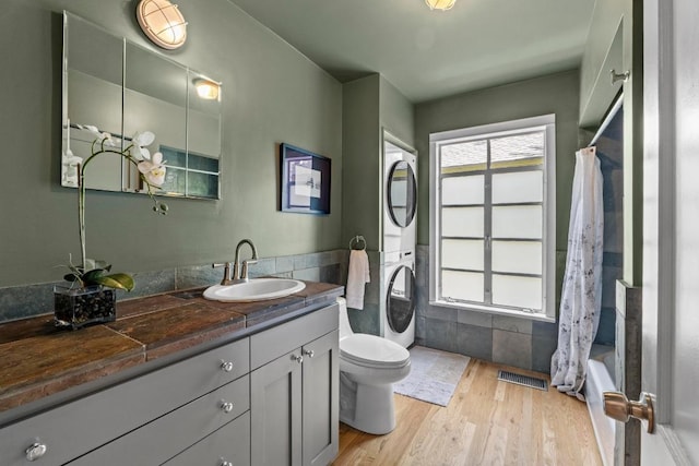 full bathroom with stacked washing machine and dryer, toilet, wood finished floors, vanity, and tile walls