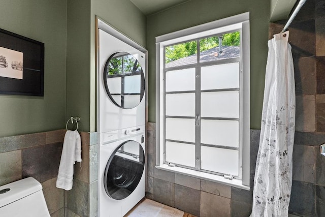laundry room featuring tile walls, stacked washer and clothes dryer, and a wainscoted wall