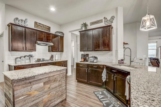 kitchen with a sink, under cabinet range hood, light stone countertops, decorative backsplash, and dark brown cabinetry