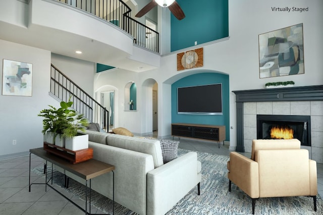 tiled living area with a high ceiling, a ceiling fan, baseboards, a tiled fireplace, and stairs