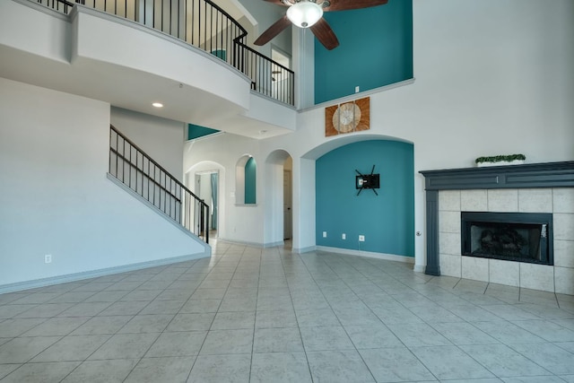 unfurnished living room with a high ceiling, ceiling fan, a tiled fireplace, tile patterned floors, and baseboards