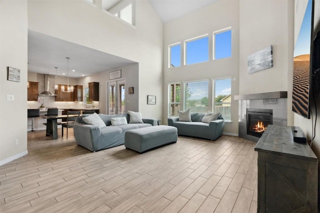 living room featuring a glass covered fireplace, light wood-type flooring, a towering ceiling, healthy amount of natural light, and baseboards