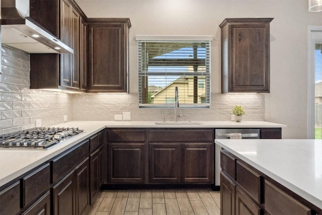 kitchen featuring wall chimney range hood, a sink, stainless steel appliances, light countertops, and dark brown cabinets