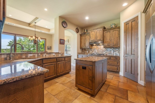 kitchen featuring a sink, backsplash, under cabinet range hood, dark stone countertops, and stainless steel refrigerator