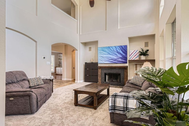 living room featuring arched walkways, a brick fireplace, a towering ceiling, carpet, and a ceiling fan