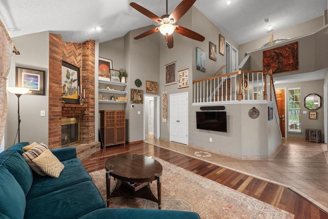 living room featuring a brick fireplace, wood finished floors, a ceiling fan, high vaulted ceiling, and stairway