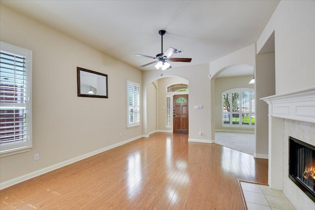 unfurnished living room featuring arched walkways, a fireplace, light wood-style floors, ceiling fan, and baseboards