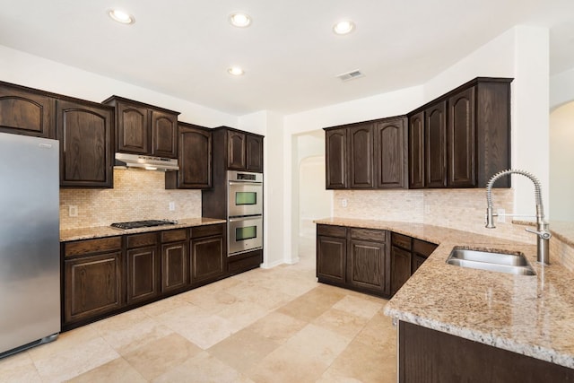 1345 Becca Teal Pl, Round Rock, TX 78681 - kitchen with appliances with stainless steel finishes, light stone countertops, dark brown cabinetry, under cabinet range hood, and backsplash