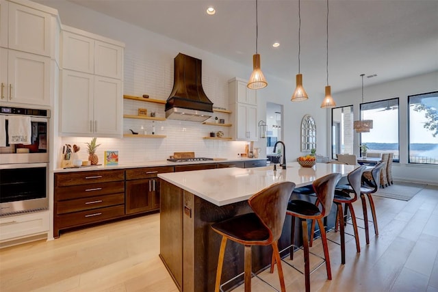 kitchen featuring appliances with stainless steel finishes, custom range hood, open shelves, light wood-style floors, and light countertops