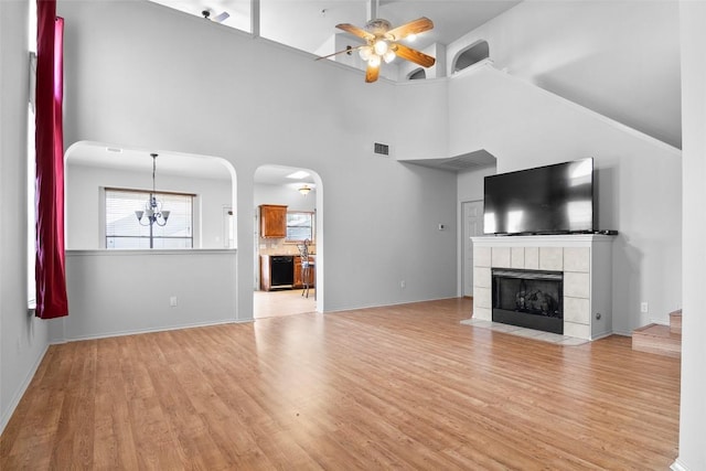 unfurnished living room featuring ceiling fan, high vaulted ceiling, light wood-style floors, a tiled fireplace, and arched walkways