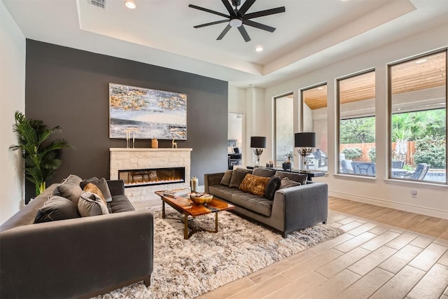 living area featuring a raised ceiling, wood finished floors, a ceiling fan, a stone fireplace, and recessed lighting