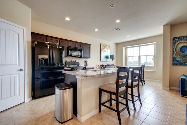 kitchen featuring black appliances, a sink, a kitchen island with sink, light stone countertops, and light tile patterned floors