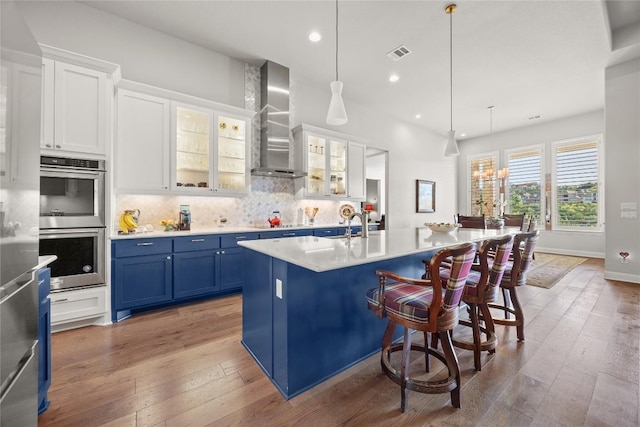 kitchen featuring wall chimney range hood, double oven, blue cabinetry, light wood-type flooring, and recessed lighting