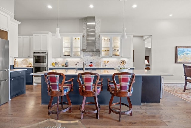 2032 Murano Ln, Leander, TX 78641 - kitchen featuring wall chimney exhaust hood, an island with sink, appliances with stainless steel finishes, wood finished floors, and light countertops