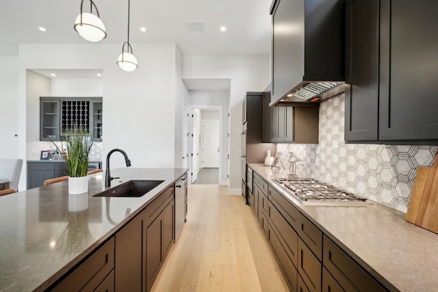 kitchen featuring a sink, wall chimney exhaust hood, backsplash, light wood-style flooring, and hanging light fixtures