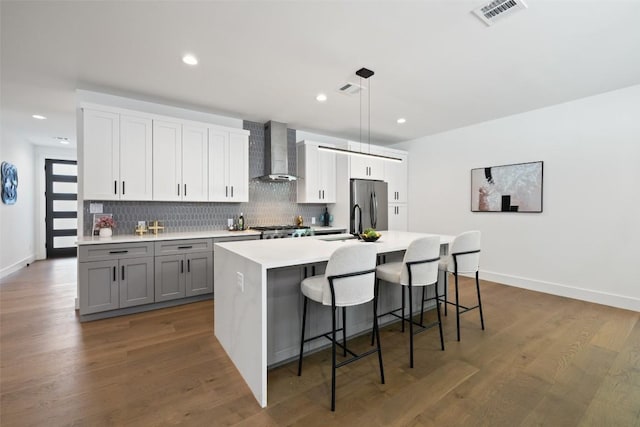kitchen featuring wall chimney range hood, freestanding refrigerator, a kitchen island with sink, backsplash, and light countertops