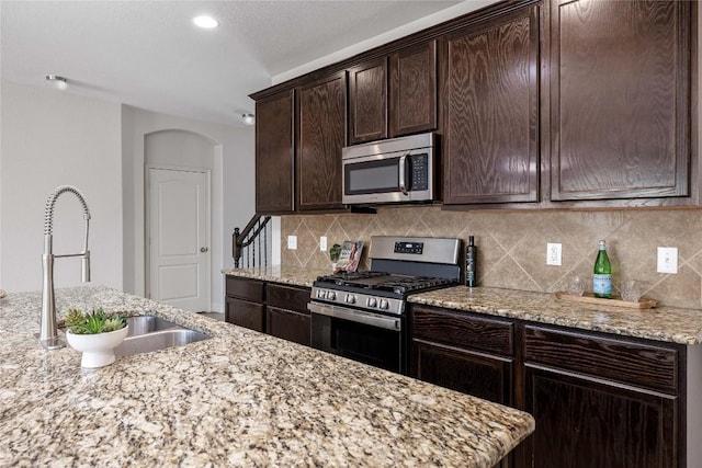 kitchen featuring stainless steel appliances, a sink, dark brown cabinets, decorative backsplash, and light stone countertops