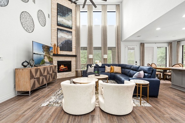 living area featuring a towering ceiling, a fireplace, ceiling fan, baseboards, and wood finished floors