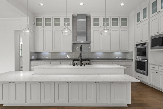 kitchen featuring wall chimney range hood, stainless steel appliances, a center island with sink, backsplash, and hanging light fixtures