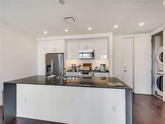 kitchen with stainless steel appliances, stacked washing machine and dryer, a sink, dark countertops, and dark wood-style flooring