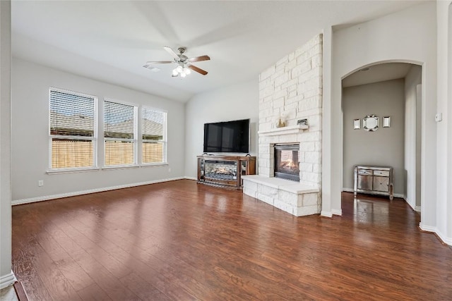 unfurnished living room featuring arched walkways, a stone fireplace, a ceiling fan, and dark wood-style flooring