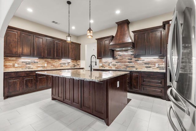 479 Brown Rd, Mcdade, TX 78650 - kitchen with freestanding refrigerator, premium range hood, a kitchen island with sink, light stone countertops, and dark brown cabinetry