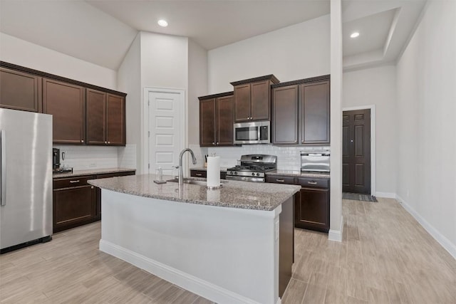 kitchen with stainless steel appliances, a sink, dark brown cabinets, a center island with sink, and tasteful backsplash
