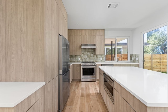 kitchen with modern cabinets, stainless steel appliances, under cabinet range hood, and light brown cabinets