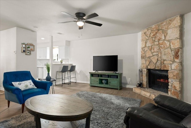 living room with ceiling fan, a stone fireplace, wood finished floors, and baseboards