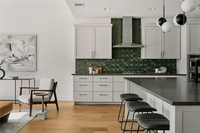 kitchen featuring wall chimney range hood, dark countertops, backsplash, stainless steel double oven, and a breakfast bar