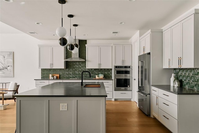 kitchen featuring stainless steel appliances, a sink, dark countertops, wall chimney range hood, and tasteful backsplash