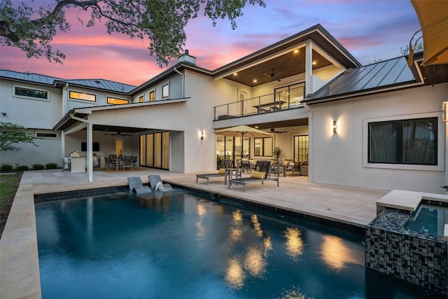 back of property at dusk featuring a balcony, a ceiling fan, a standing seam roof, and stucco siding