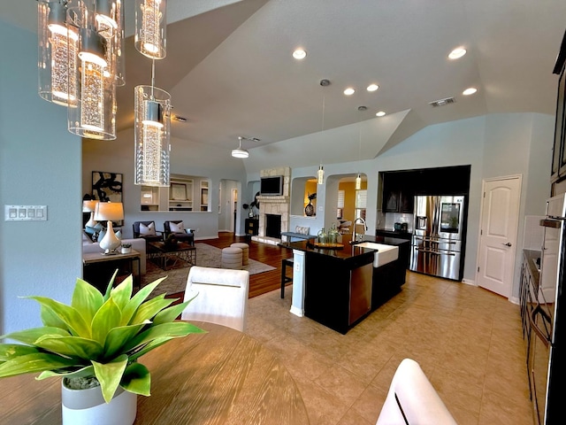 kitchen with arched walkways, a sink, stainless steel fridge with ice dispenser, dark countertops, and a kitchen island with sink