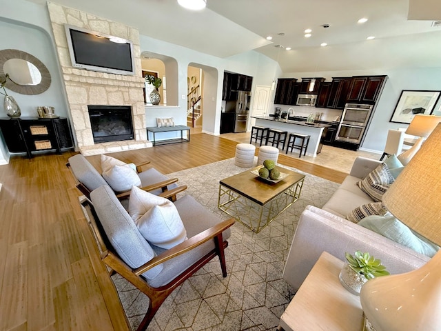living area featuring arched walkways, lofted ceiling, light wood-style flooring, a stone fireplace, and baseboards