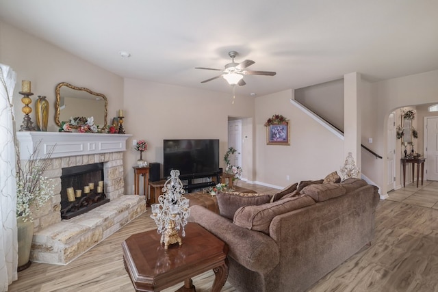 living room featuring ceiling fan, stairs, light wood-style floors, a fireplace with raised hearth, and baseboards